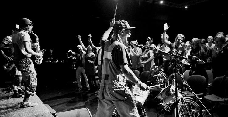 Bicycle Beat on stage: the drummer on the bike with a stick raised, a saxophonist beside him and a crowd with their hands up